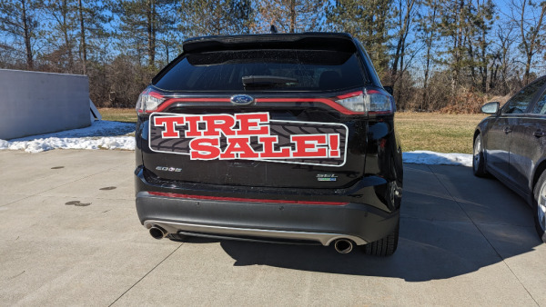 A small reusable magnetic sign advertises a tire sale on the hatchback of an SUV
